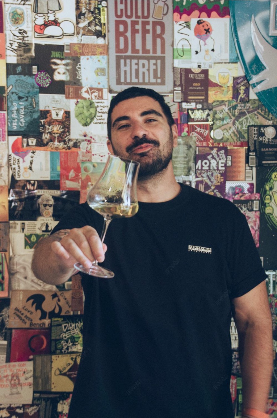 Matteo con un calice in mano davanti a una parete di adesivi di birrifici